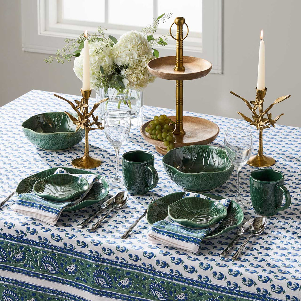 Elegant table setting with green ceramic dishes, gold candle holders, and white flowers on a patterned tablecloth.