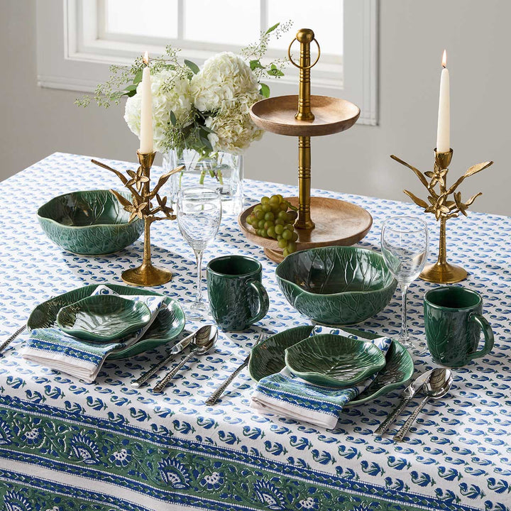 Elegant table setting with green ceramic dishes, gold candle holders, and white flowers on a patterned tablecloth.