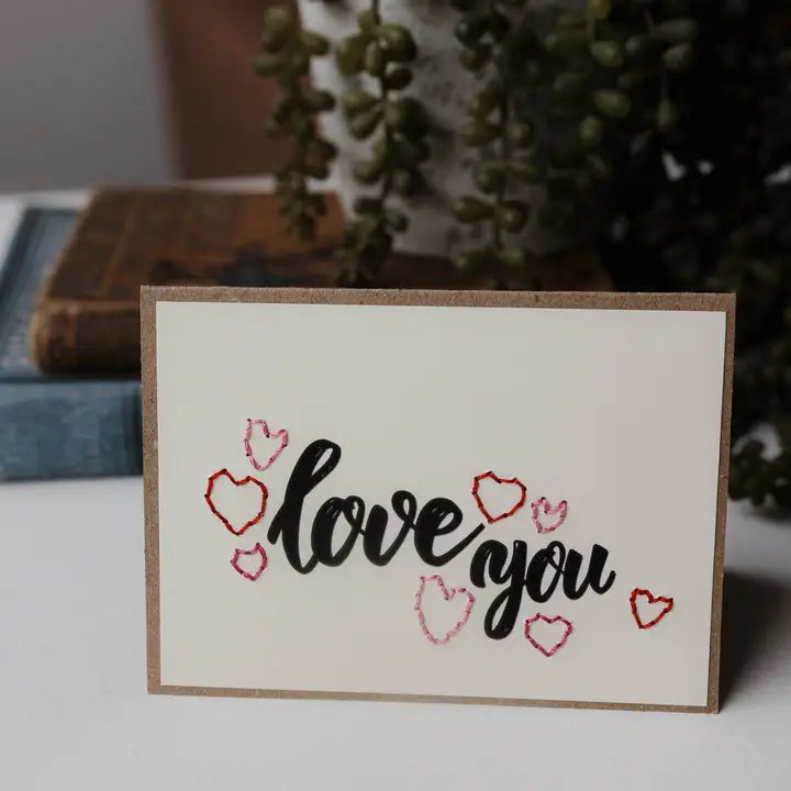 A handmade greeting card with 'love you' written in black script and surrounded by small red and pink heart outlines, set against a blurred background of books and a green trailing plant.
