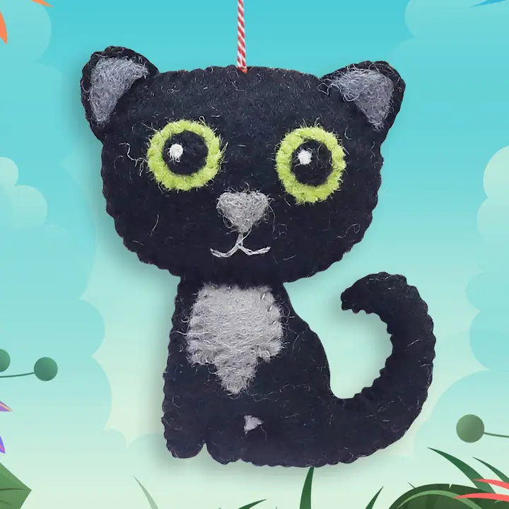 A black felt cat ornament with bright lime green eyes, a gray chest and ears, and a red-and-white hanging string, shown against a light blue illustrated background.