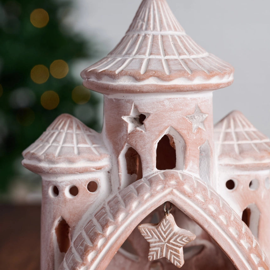 A close-up of a decorative castle-shaped ceramic ornament or figurine in a pale pink and off-white glaze, with star and star cutouts and a hanging star ornament, against a softly blurred holiday background.