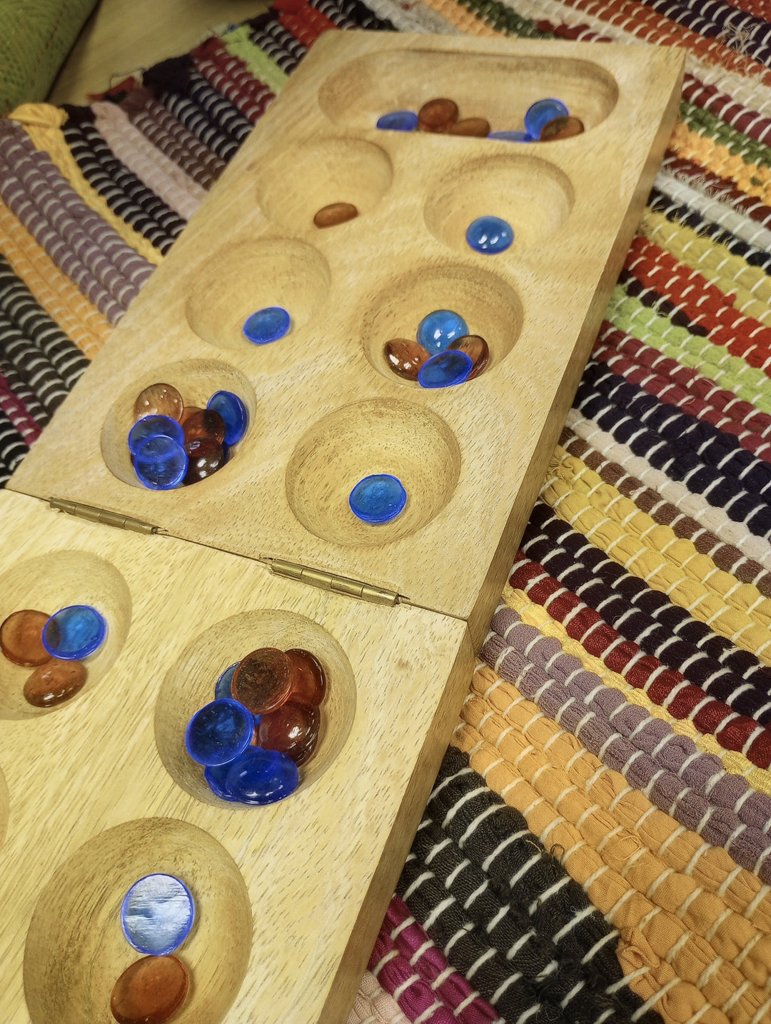 A wooden Mancala game board, shown open and filled with blue and orange playing pieces, placed on a colorful woven textile background.