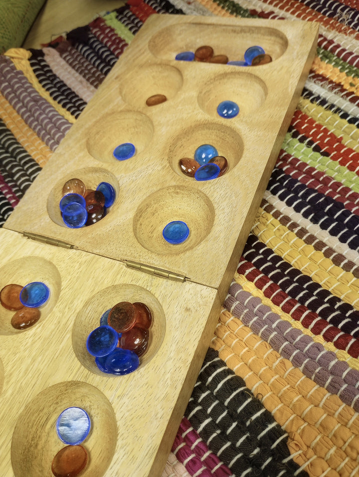 A wooden Mancala game board, shown open and filled with blue and orange playing pieces, placed on a colorful woven textile background.