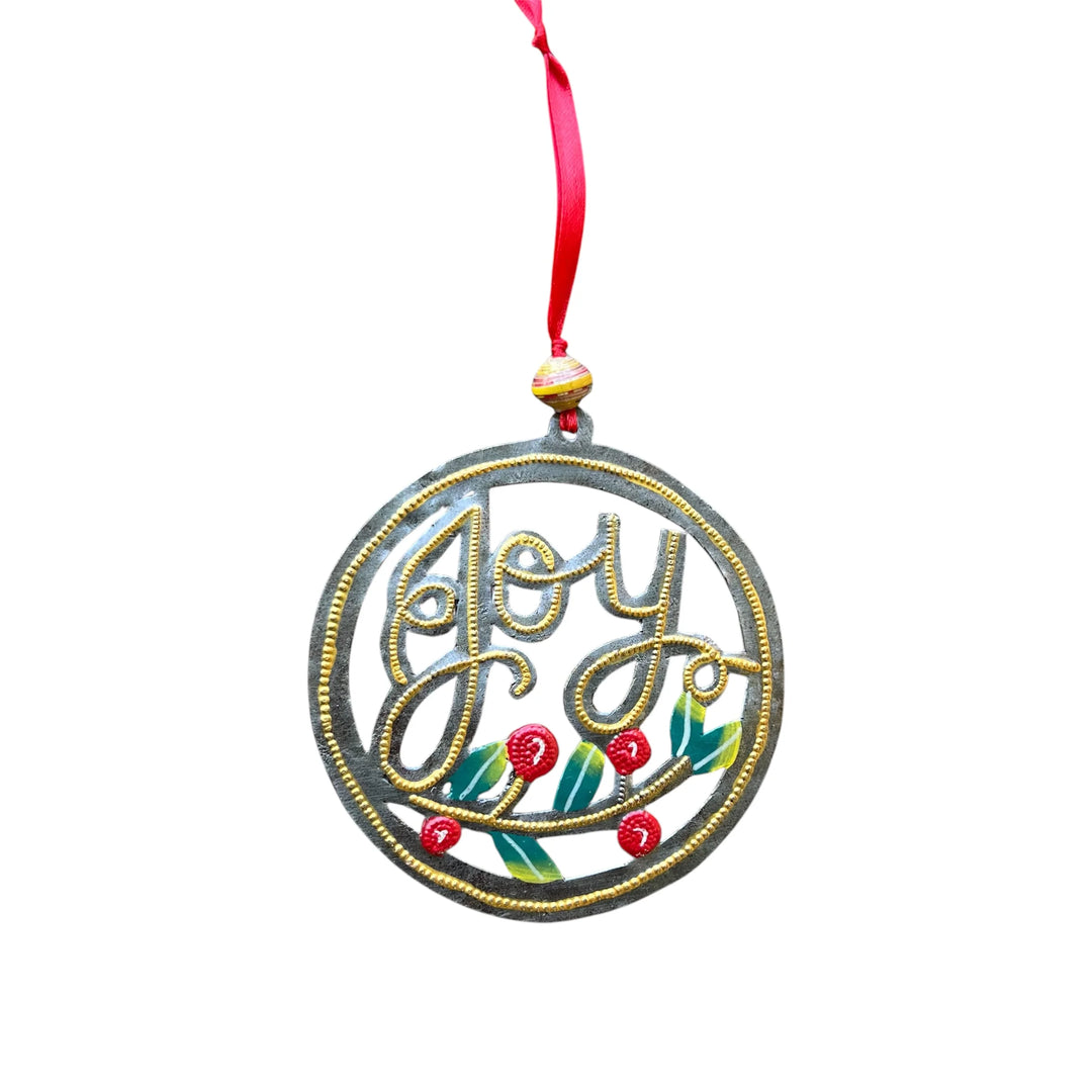 A round metal Christmas ornament with the word 'Joy' in gold cursive lettering, decorated with red berries and green leaves, hanging from a red ribbon.