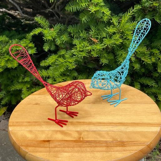 Two metal wire birds, blue and red, on a table outside by a tree.