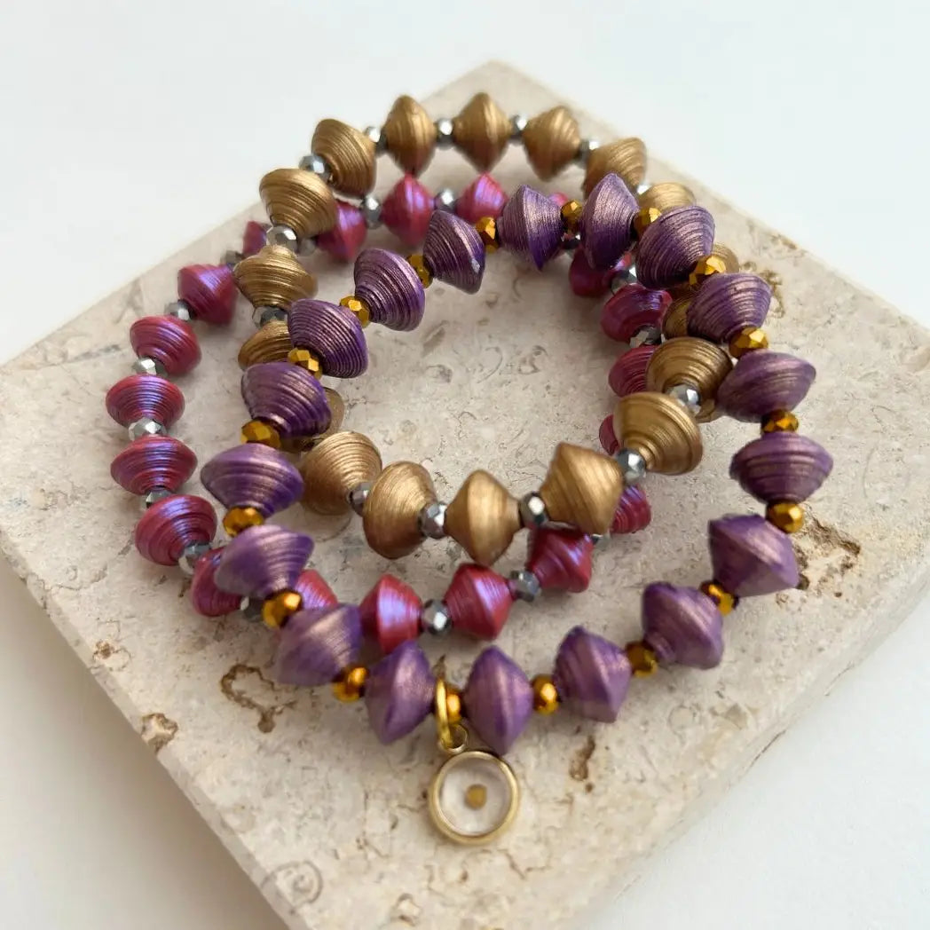 A set of beaded stretch bracelets with purple and metallic gold spiral beads, displayed on a textured beige stone tile.
