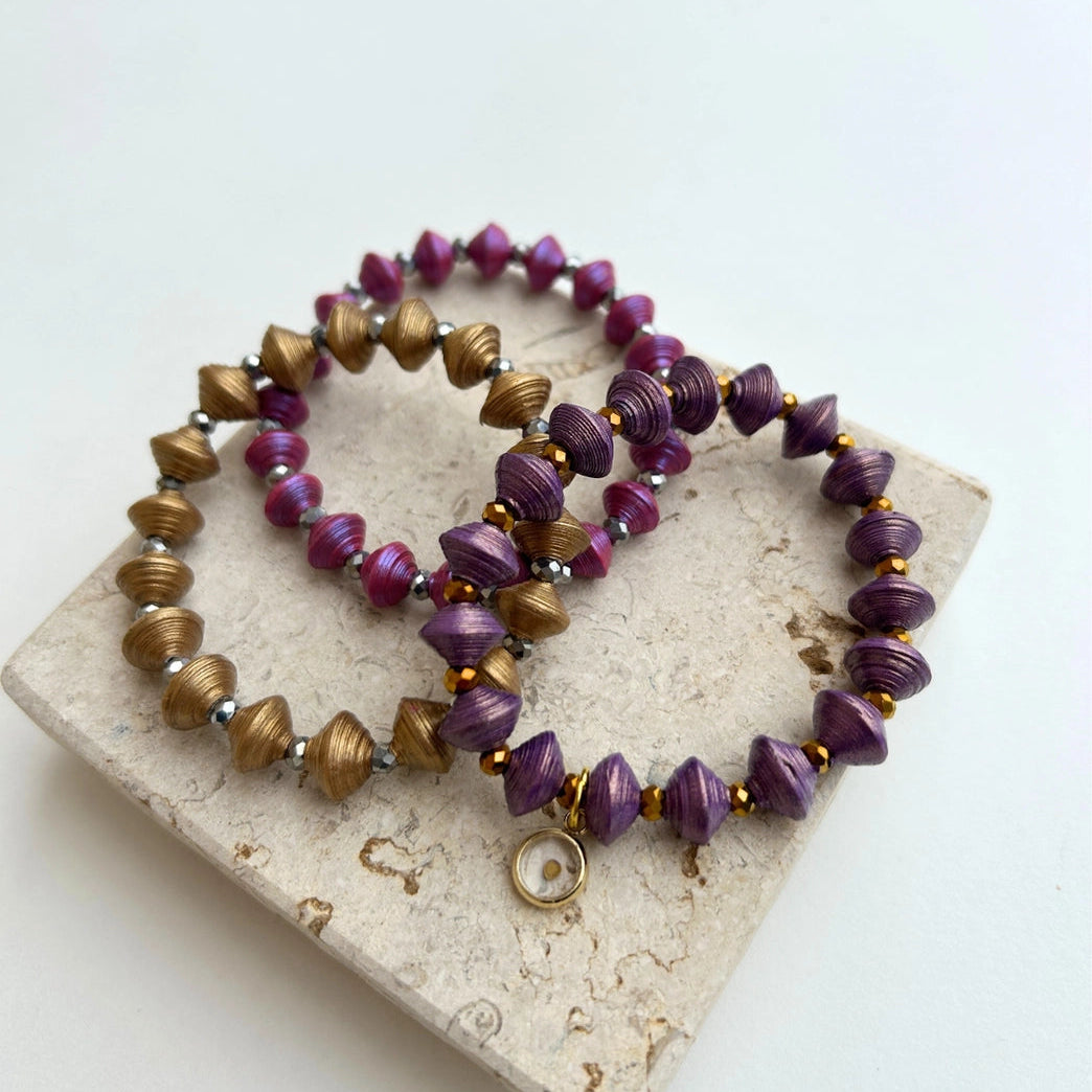 Two beaded stretch bracelets with purple and metallic bronze spiral beads, displayed on a textured stone tile against a white background.