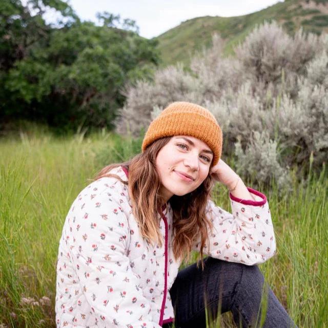 Person wearing a mustard yellow beanie and floral jacket sitting in a grassy field with trees in the background