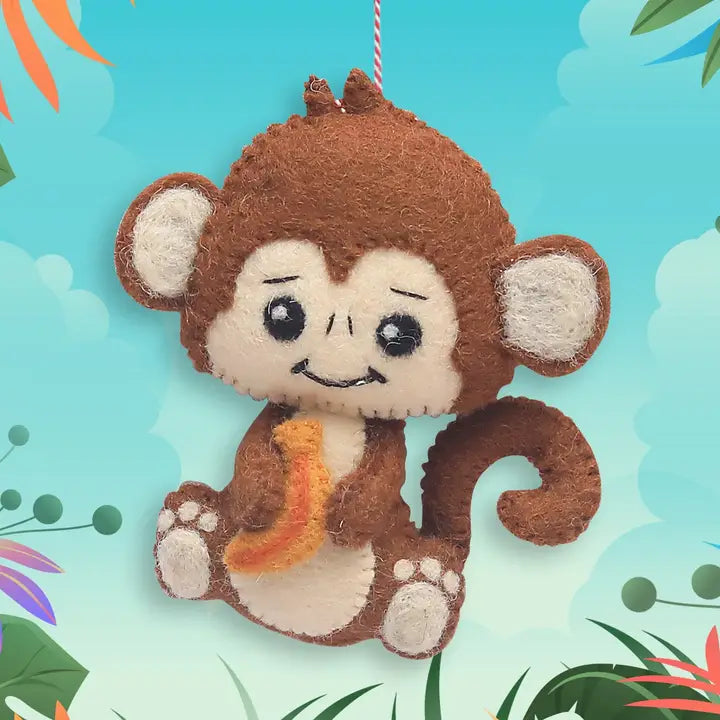 A brown and cream felt monkey hanging ornament holding a yellow banana, set against a tropical-themed light blue background with leaves and clouds.