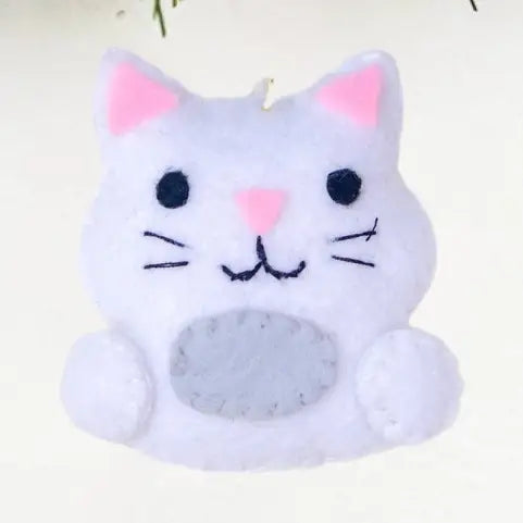 A white felt cat-shaped ornament or decoration with pink inner ears, black embroidered eyes and whiskers, a pink stitched nose, and a gray oval patch on the belly, shown against a plain light background.
