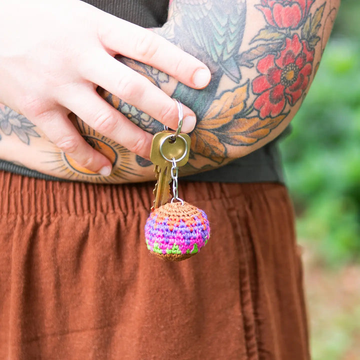 A close-up lifestyle shot of a person's tattooed arm holding a small, colorful crocheted hacky sack keychain charm with keys, wearing a brown skirt.