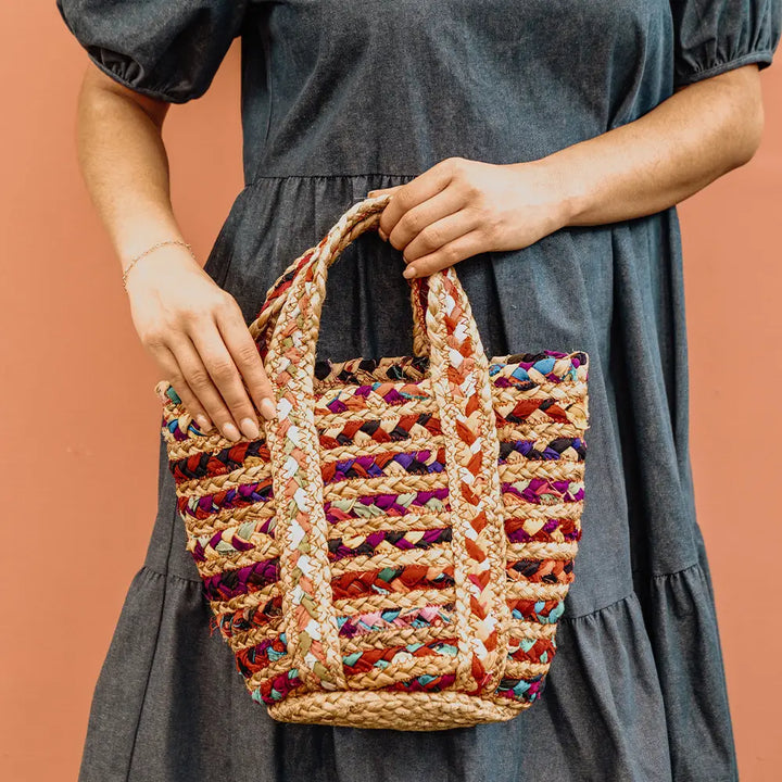 A person in a denim dress holds a woven straw tote bag with colorful striped fabric accents and two top handles, set against a solid terracotta background.