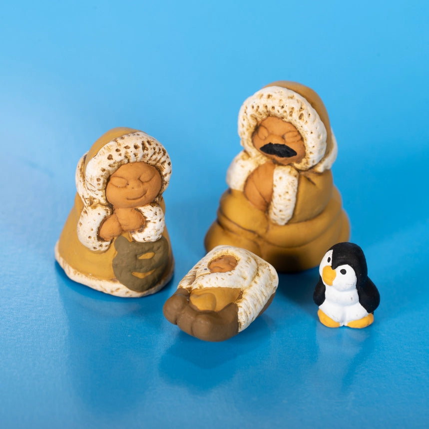 Three small clay figurines on a blue background: a seated woman, a seated man, and a baby in a manger, plus a small penguin figurine.