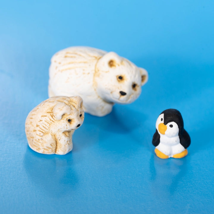 Three small figurines on a bright blue background: a large cream-colored polar bear, a smaller cream-colored cub, and a black-and-white penguin with yellow accents.