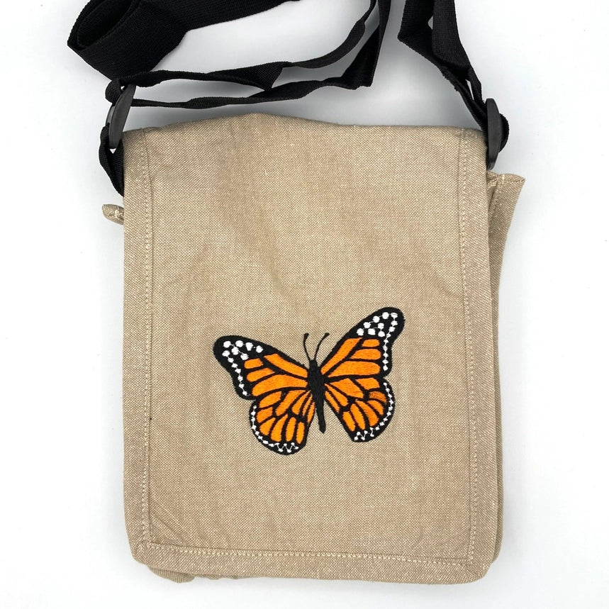 A beige canvas shoulder bag with a black adjustable strap, featuring an embroidered orange and black butterfly on the front, set against a plain white background.
