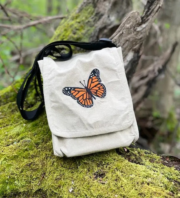 A small off-white canvas pouch bag with a black strap, featuring an embroidered orange and black monarch butterfly on the front, resting on a mossy tree branch outdoors.