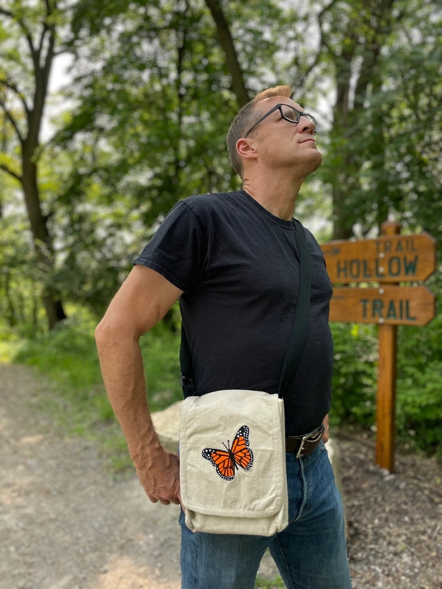 Man standing outdoors with a butterfly-themed bag, looking up at trees.