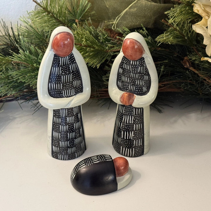 Two soapston nativity figures with patterned clothing and a third figure on a white surface with greenery in the background.
