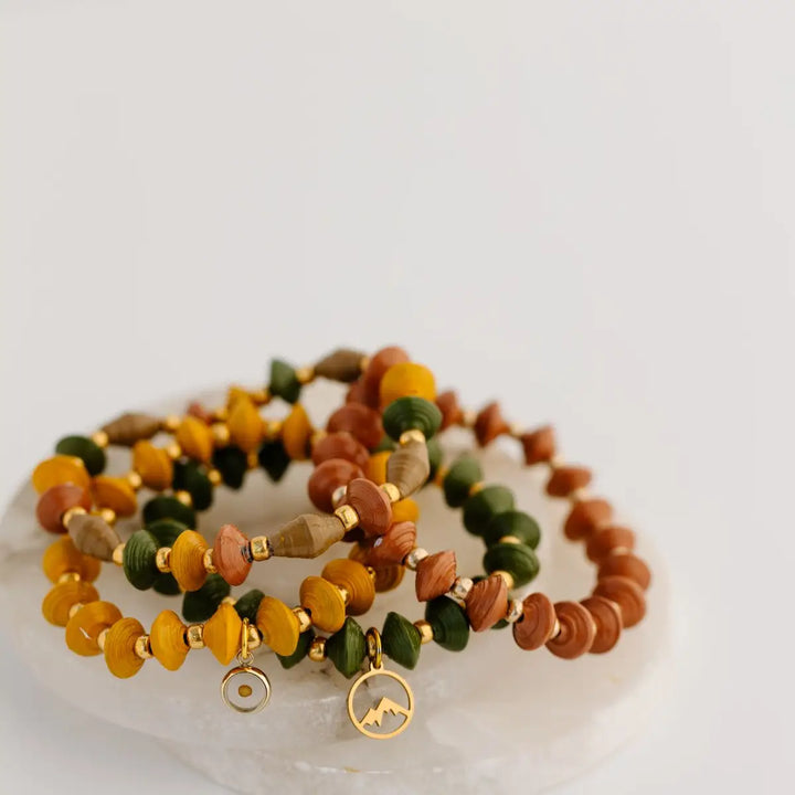 Three beaded stretch bracelets with gold charms, displayed on a white stone slab against a light gray background. The bracelets feature natural wood beads in yellow, orange, green, and brown, separated by small gold spacer beads.