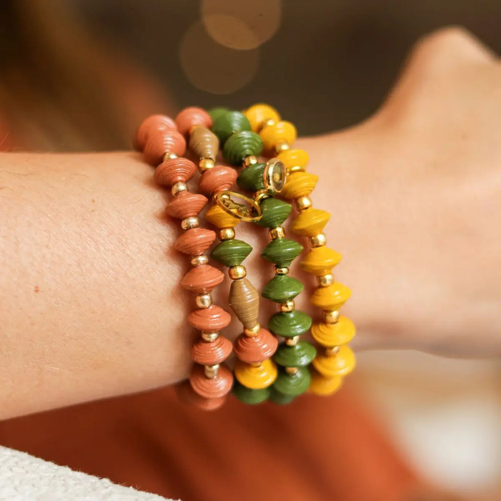 A close-up of a person's wrist wearing three beaded wrap bracelets in terracotta, olive green, and mustard yellow with small gold spacer beads and a butterfly charm.