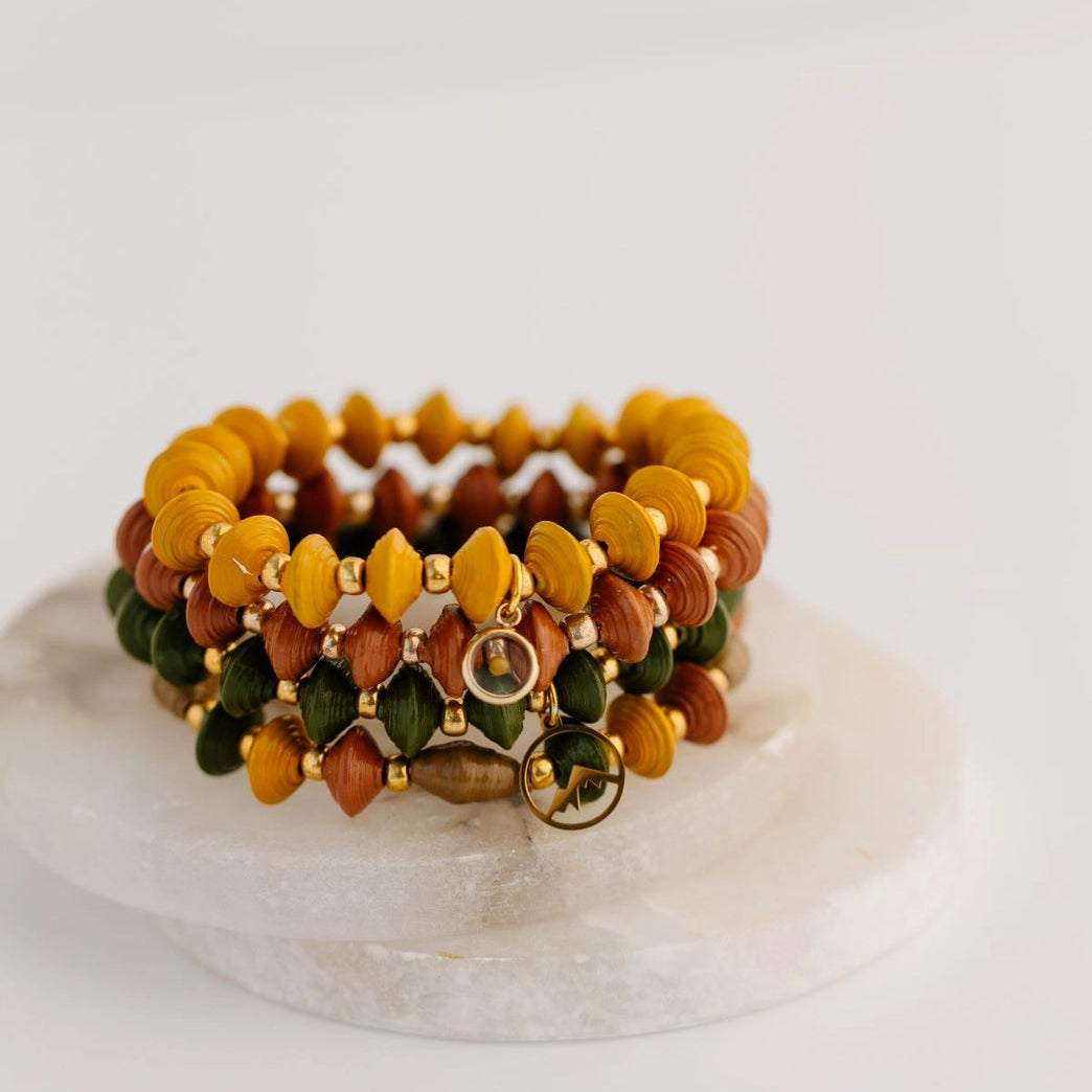 A stack of three beaded stretch bracelets displayed on a round white stone tray against a clean white background. The bracelets feature faceted beads in mustard yellow, terracotta orange-brown, and olive green, separated by small gold spacer beads, with a gold-toned clasp.