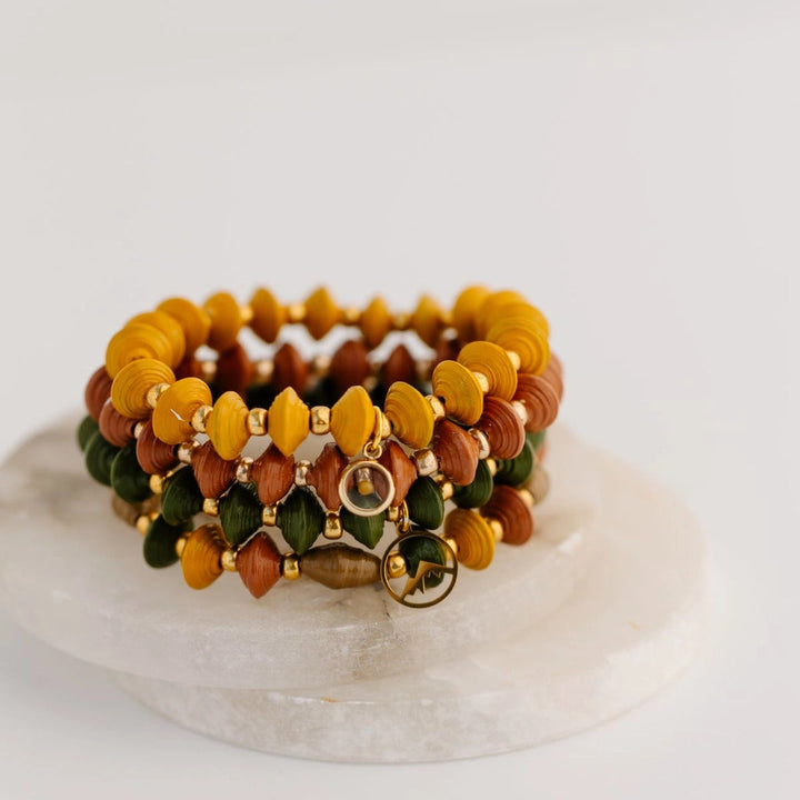 A stack of three beaded stretch bracelets displayed on a round white stone tray against a clean white background. The bracelets feature faceted beads in mustard yellow, terracotta orange-brown, and olive green, separated by small gold spacer beads, with a gold-toned clasp.