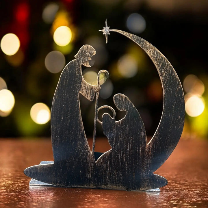 A dark metal cutout nativity candleholder decoration on a wooden surface, featuring Mary, Joseph, baby Jesus, a crescent moon, and a star, with a blurred festive background of warm lights.