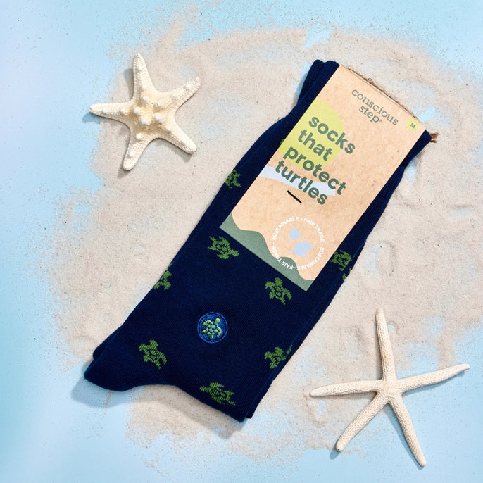Navy blue socks with green turtle patterns on a sand and starfish background, featuring 'Conscious Step' packaging.