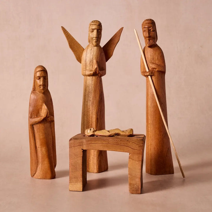 A set of hand-carved wooden figurines depicting religious figures, including an angel with outstretched wings and two robed figures, arranged around a small wooden table with a carved piece on top, photographed against a solid warm beige background.