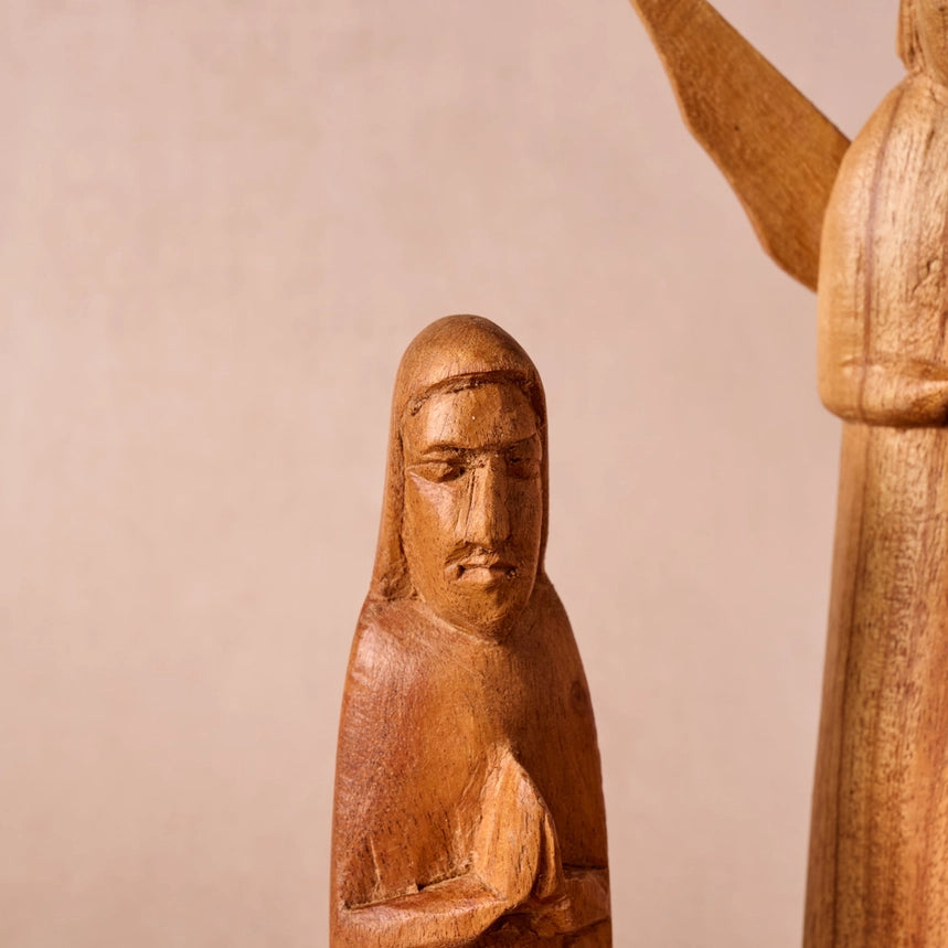 Close-up of a carved wooden figurine with a human face and draped robes, shown against a plain beige background with part of another wooden statue visible on the right.