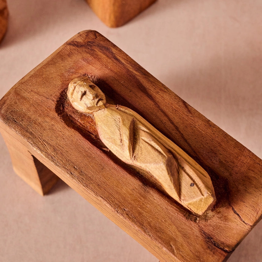 A hand-carved wooden figurine of a reclining figure, mounted on a rustic rectangular wooden plaque, set against a neutral beige background.