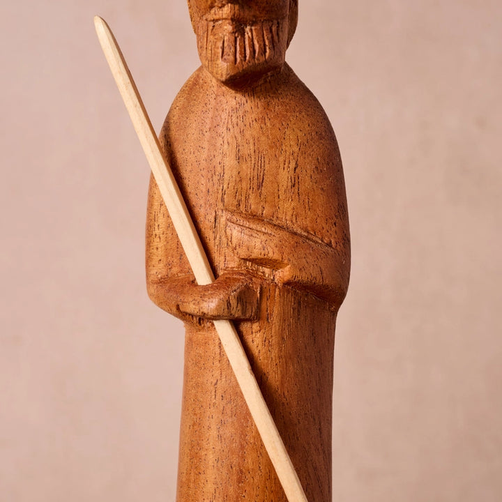 A close-up of a hand-carved wooden figurine of a robed man holding a staff, shown against a plain beige background.