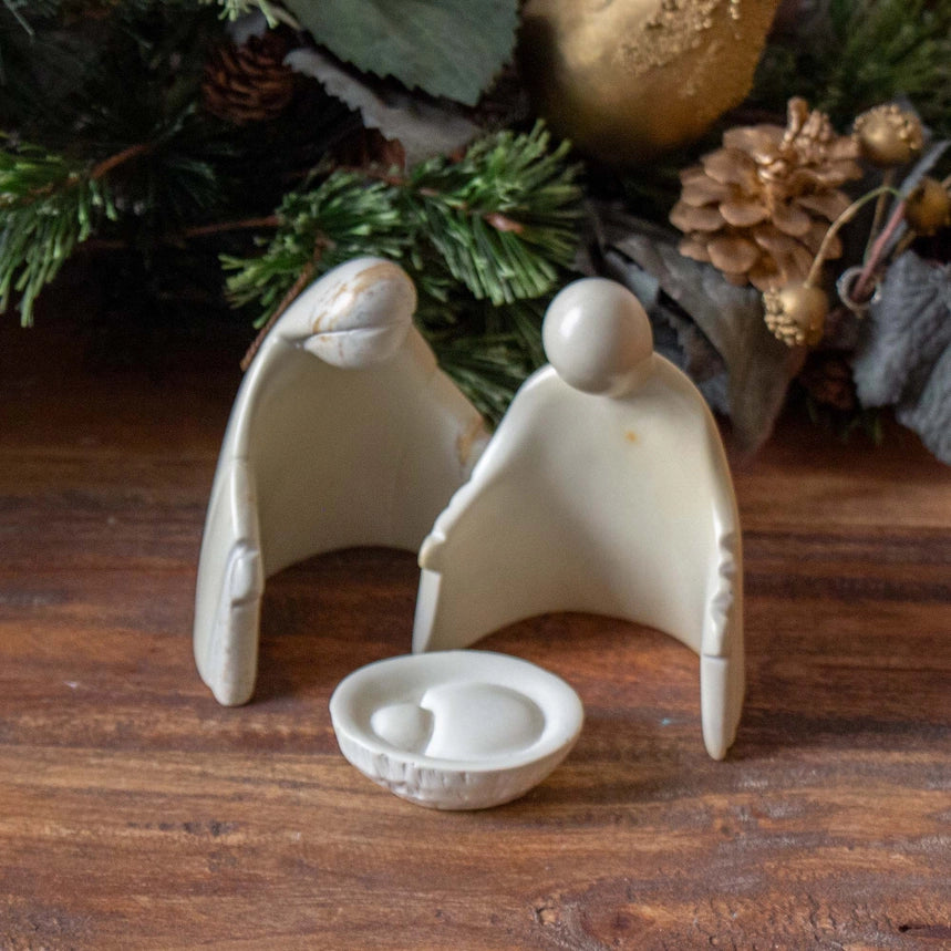 A small ceramic nativity scene with two angel figures and a shallow basket holding Baby Jesus, displayed on a wooden surface with pine branches and holiday ornaments in the background.