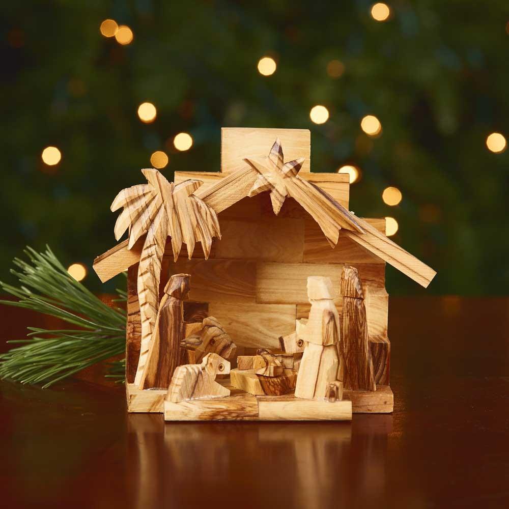 Wooden nativity scene with a palm tree and stable against a blurred Christmas tree background.