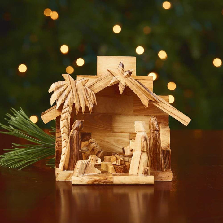 Wooden nativity scene with a palm tree and stable against a blurred Christmas tree background.