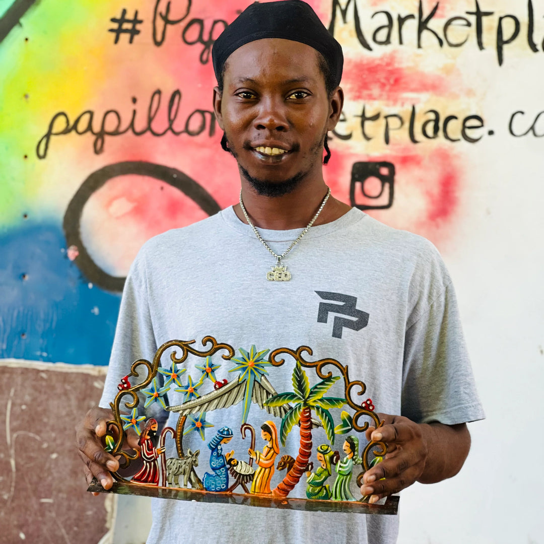 A man wearing a black cap and a light gray t-shirt with a graphic print holds up a colorful hand-painted nativity scene plaque in front of a graffiti-covered wall.