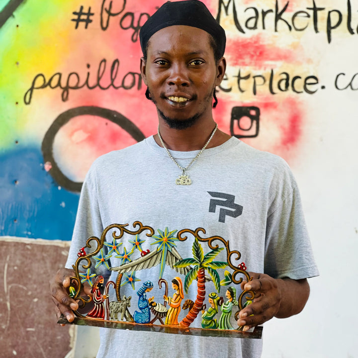 A man wearing a black cap and a light gray t-shirt with a graphic print holds up a colorful hand-painted nativity scene plaque in front of a graffiti-covered wall.