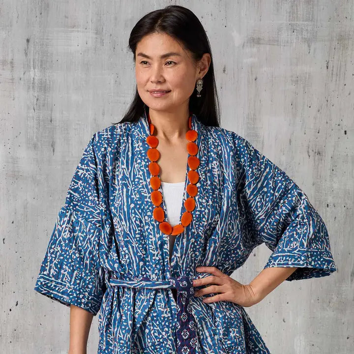 Woman wearing a blue patterned kimono with an orange tagua necklace against a gray concrete wall.