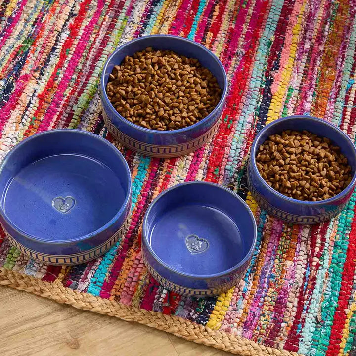 Four ceramic pet bowls in blue and white are arranged on a colorful woven rug, with two bowls filled with kibble. The set includes one large shallow bowl, one medium shallow bowl, and two deeper bowls, all with a small heart design in the center.