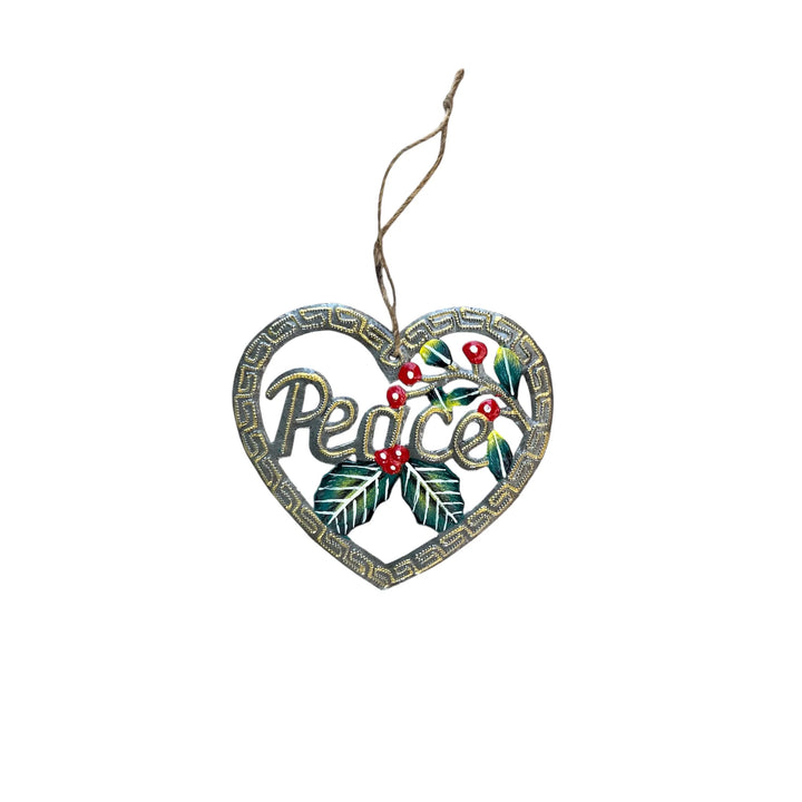 A heart-shaped metal Christmas ornament featuring the word 'Peace' in cursive, decorated with green holly leaves, red berries, and a Greek key border, hanging from a twine string against a white background.