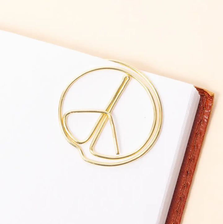 A gold metal peace sign bookmark resting on the corner of a white notebook, with a brown textured book edge visible on the right against a warm cream background.