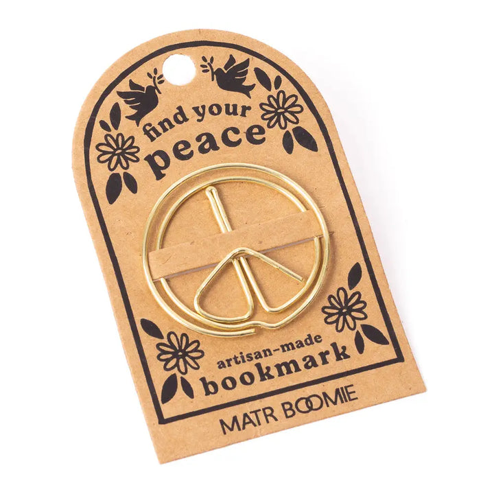 A gold metal bookmark shaped like a peace sign, attached to a brown kraft paper display card with black printed text and illustrations. The card reads 'find your peace', 'artisan-made bookmark', and 'MATR BOOMIE'.