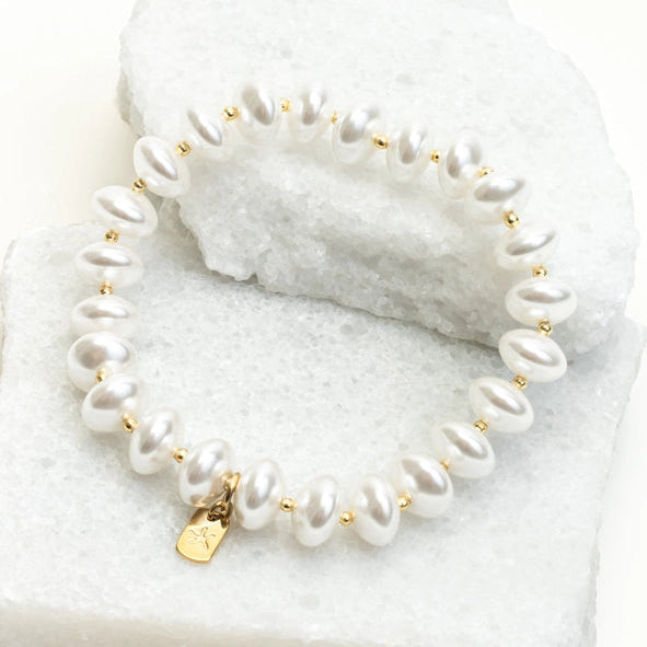 A stretch bracelet made of white baroque pearls separated by small gold spacer beads, with a small gold heart charm, displayed on a white textured stone block.