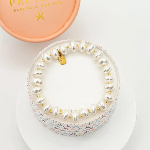 A Starfish Project pearl bracelet displayed on a round white stand, with its peach-colored branded lid placed above on a clean white background.