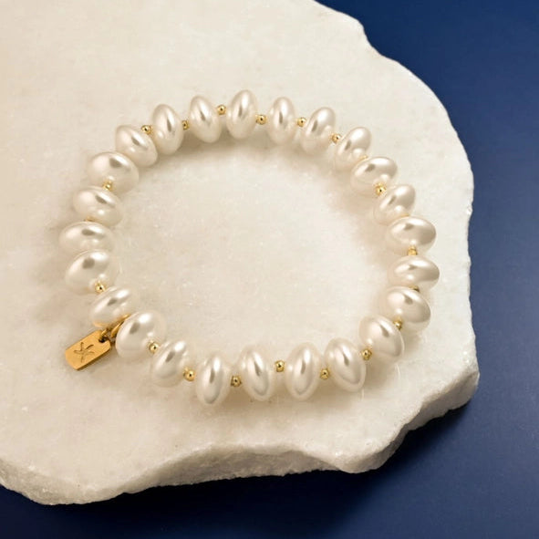 A pearl beaded bracelet with small gold spacer beads and a gold charm, displayed on a white textured stone against a dark blue background.