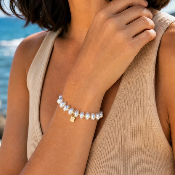 A close-up lifestyle shot of a woman on a beach wearing a delicate white pearl beaded bracelet with a small gold square charm.