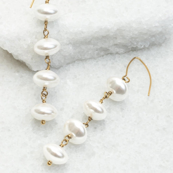 A pair of gold-tone drop earrings with irregular white freshwater pearls, displayed on a textured white stone surface.
