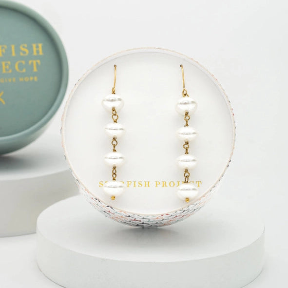White pearl drop earrings with gold hooks, displayed on a white circular stand with a white pedestal base. A mint green round box with gold lettering is partially visible on the left.
