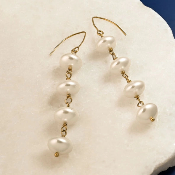 A pair of gold dangle earrings with irregular white freshwater pearls, displayed on a white textured stone slab against a dark blue background.