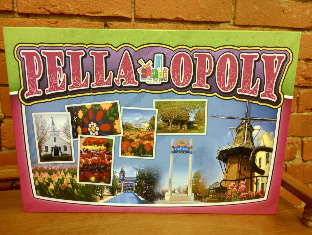 A board game box with 'PELLA-OPOLY' written on it, featuring images of Pella, Iowa, including a windmill, tulips, and a corn maze.